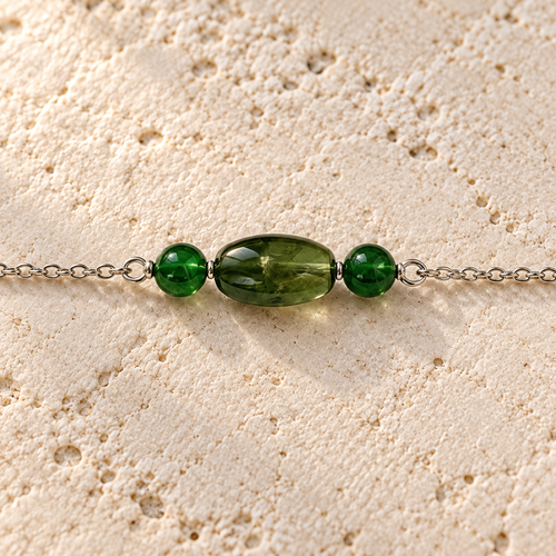 Close-up of a dark green oval bead between two small green beads on a silver chain, resting on ivory stone.